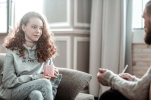 a young woman holds a coffee mug and sits on a couch and listens to her therapist explain the benefits of residential eating disorder treatment