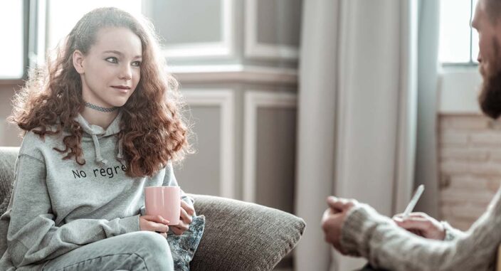 a young woman holds a coffee mug and sits on a couch and listens to her therapist explain the benefits of residential eating disorder treatment