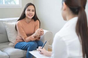 a young woman patient sits in her therapist's office discussing binge eating disorder while her therapist takes notes on her clipboard