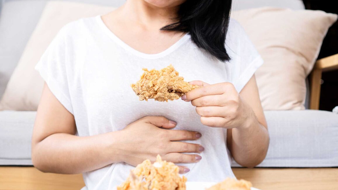 a woman holds one hand to her stomach while the other one holds a chicken drumstick and realizes how binge eating affects her body