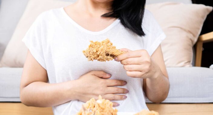 a woman holds one hand to her stomach while the other one holds a chicken drumstick and realizes how binge eating affects her body
