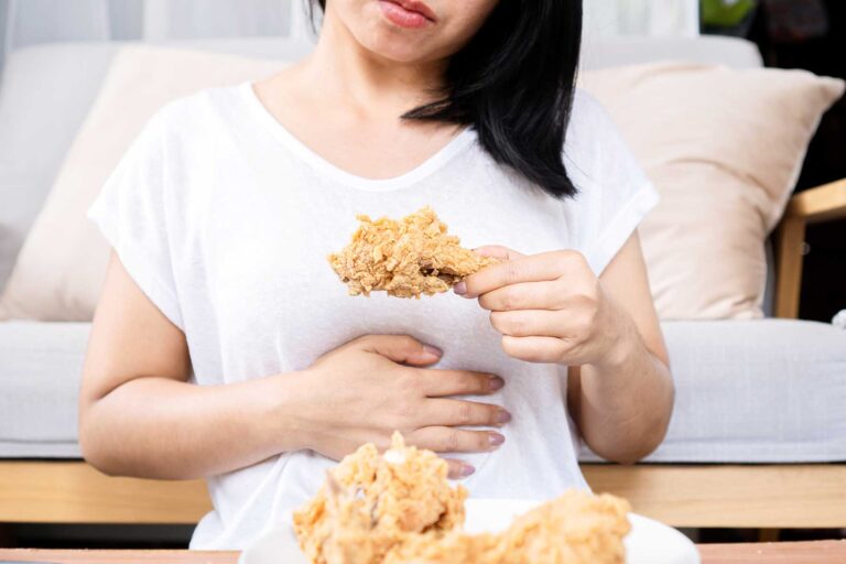 a woman holds one hand to her stomach while the other one holds a chicken drumstick and realizes how binge eating affects her body