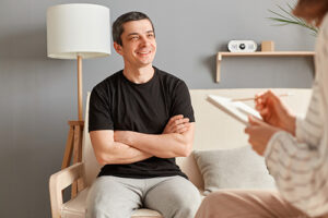 man sitting with his arms crossed smiles and listens to his therapist who is taking notes about the best options for addiction treatments for him