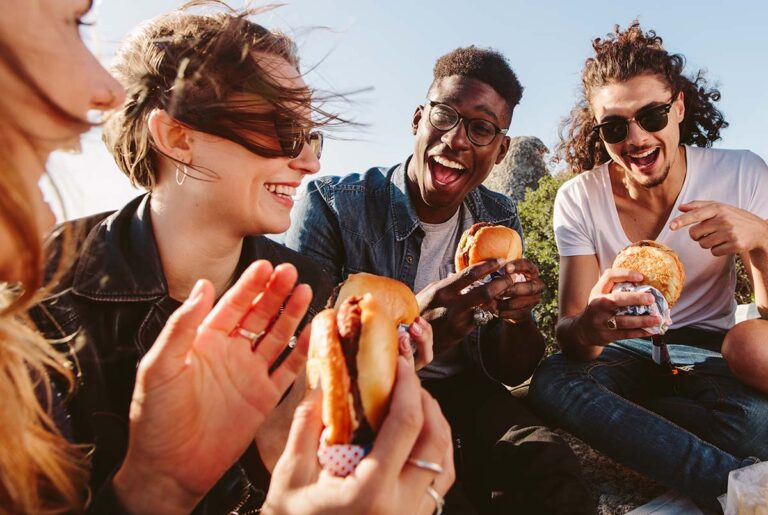 four friends are outside eating breakfast sandwiches and smiling while discussing how they might help their friend with an eating disorder