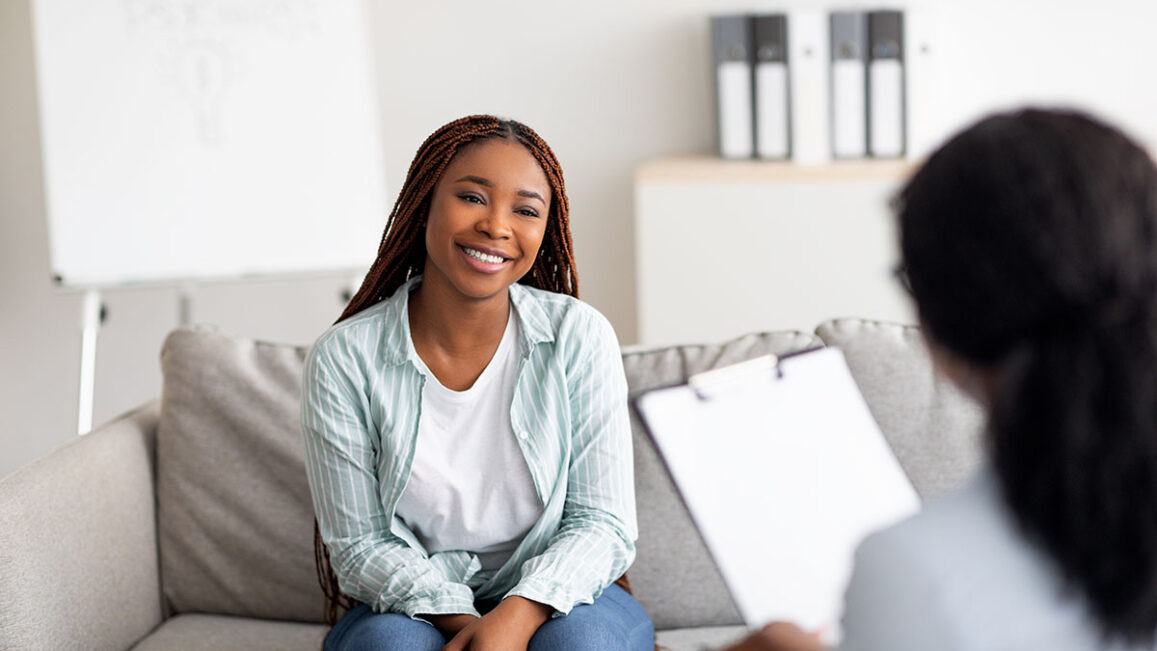 a smiling woman sits on a couch while talking to her therapist about the differences of dysphoria vs dysmorphia