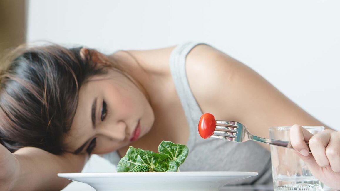 woman holds a fork with a cherry tomato while resting her head on the table next to her plate and asks herself what is refeeding syndrome