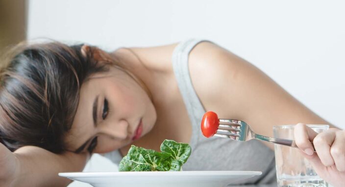 woman holds a fork with a cherry tomato while resting her head on the table next to her plate and asks herself what is refeeding syndrome