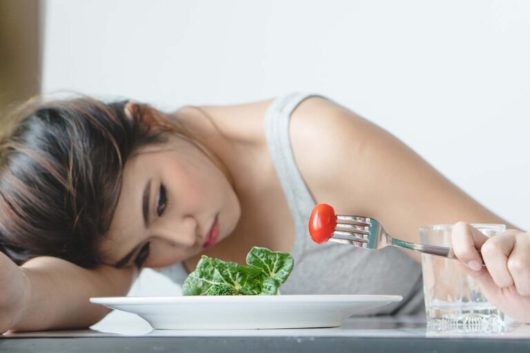 woman holds a fork with a cherry tomato while resting her head on the table next to her plate and asks herself what is refeeding syndrome