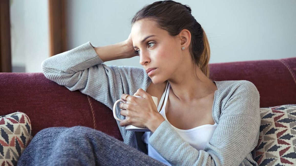 woman holding a coffee mug and sitting on a couch looking into distance wonders is anxiety genetic