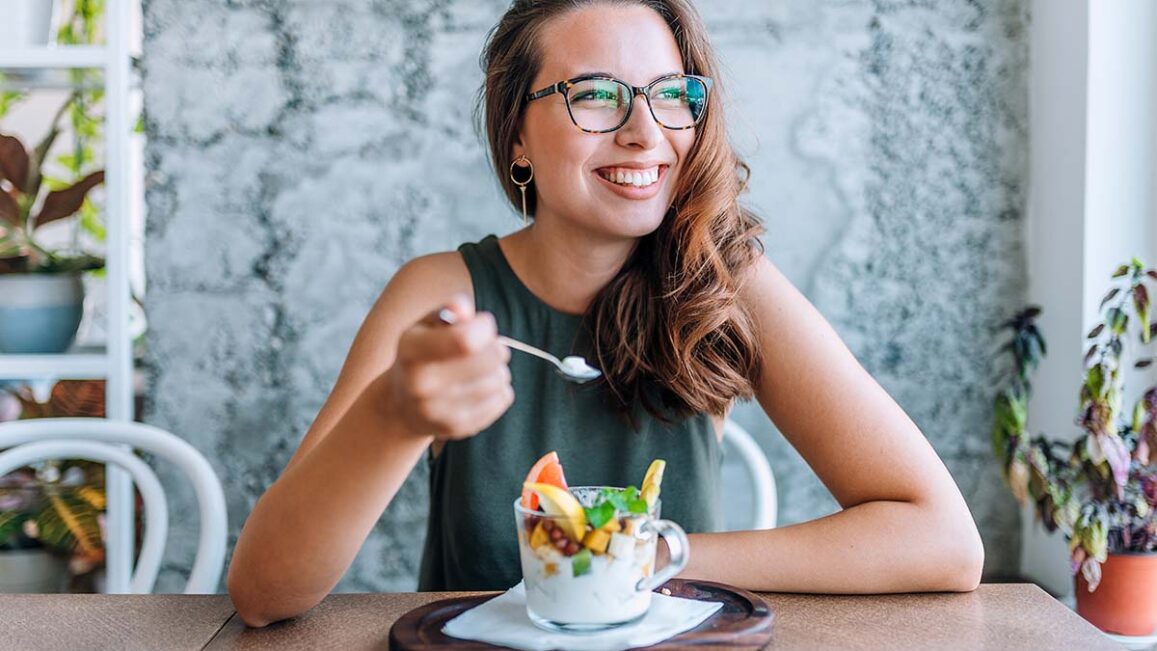 a woman wearing glasses holding a spoon while eating a parfait wonders about the long-term effects of not eating enough